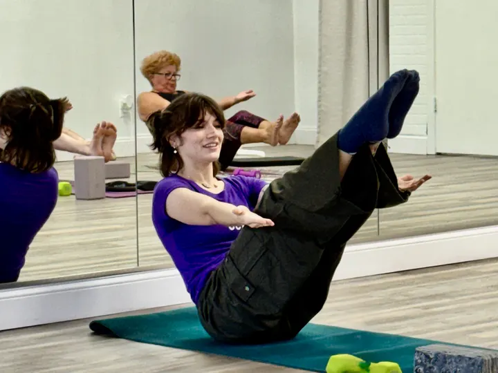 A member smiling as she holds a Pilates boat pose on her mat
