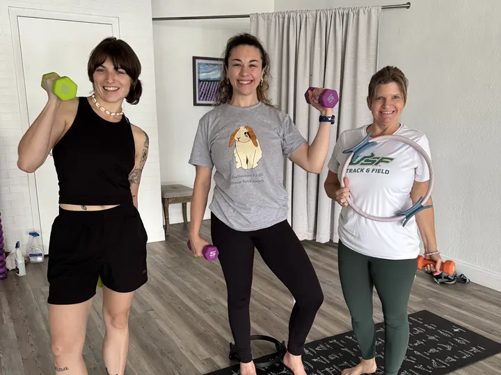 Three members smiling in the studio holding light hand weights and a Pilates ring