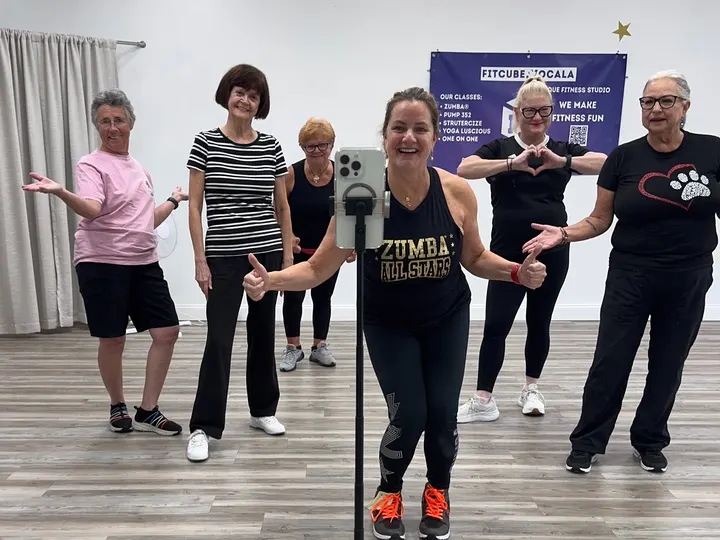 Lauren in a Zumba All Stars tank giving a thumbs up, with smiling members behind her in the studio
